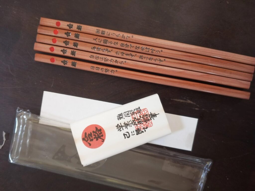 Five wooden Japanese good luck pencils with red dots near their erasers and motivational Japanese text printed on their barrels (reading "It's difficult," "Don't rely on others. If you do it yourself, it will come true," "Don't be in a hurry, don't be in a hurry," "Self-contained accommodation is from effort," and "Your own way of work?"), displayed above their white paper wrapper featuring a red circular logo and Japanese calligraphy, all arranged on a dark surface with a clear plastic case visible at the bottom of the frame.