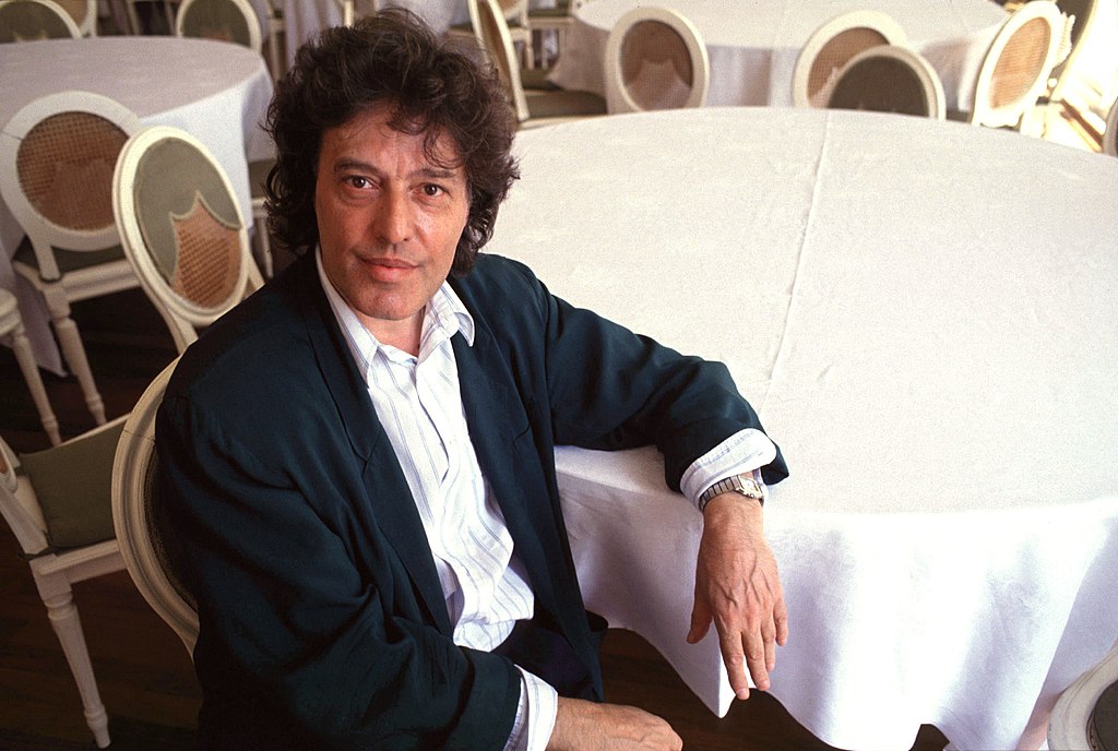 Tom Stoppard, wearing a black jacket over a white dress shirt, elbow on a round table, in 1990 at the Venice Film Festival. CC-BY-SA licensed photo by Gorup de Besanez